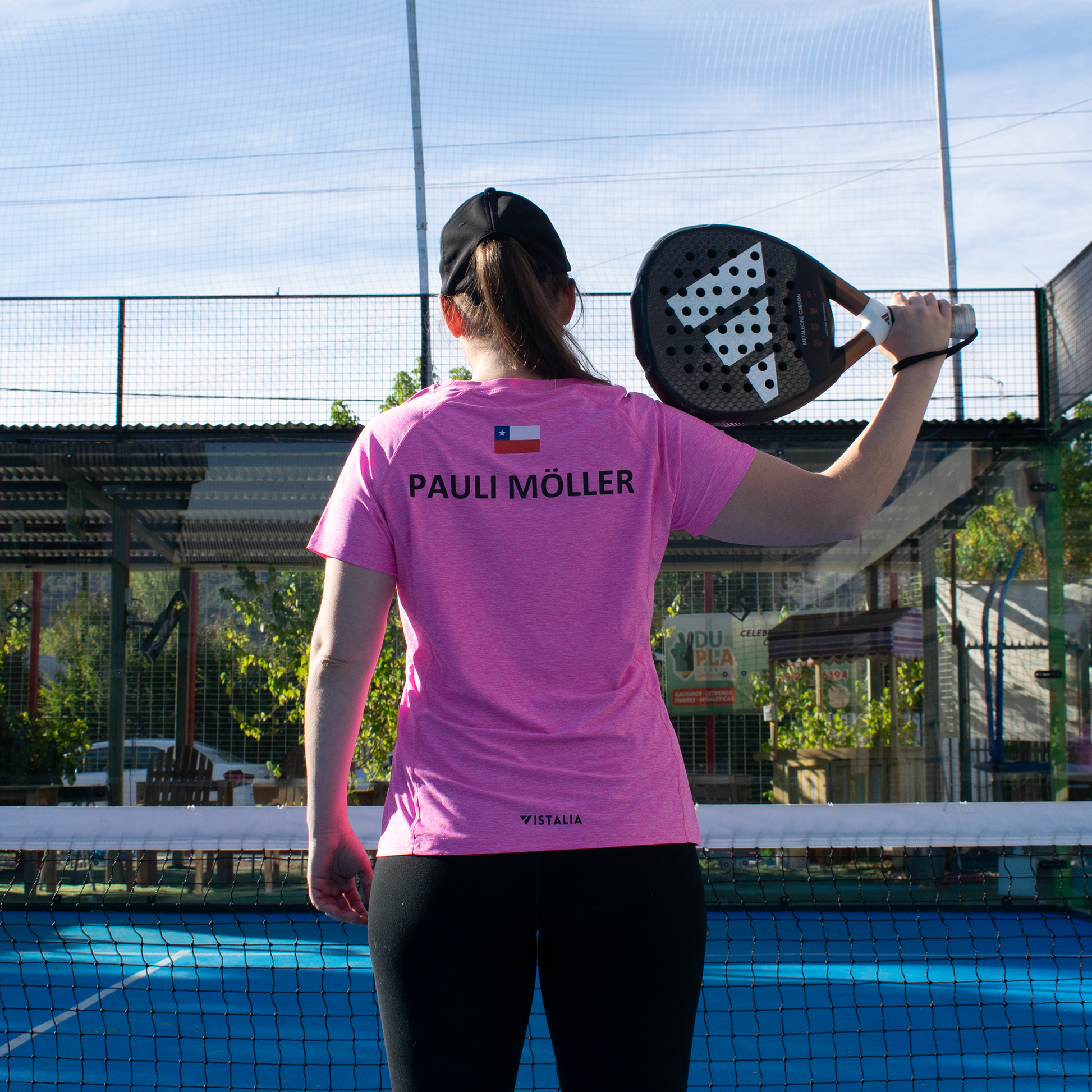 mujer de espalda en red de cancha de pádel apoyando su pala con su mano derecha en su hombro derecho