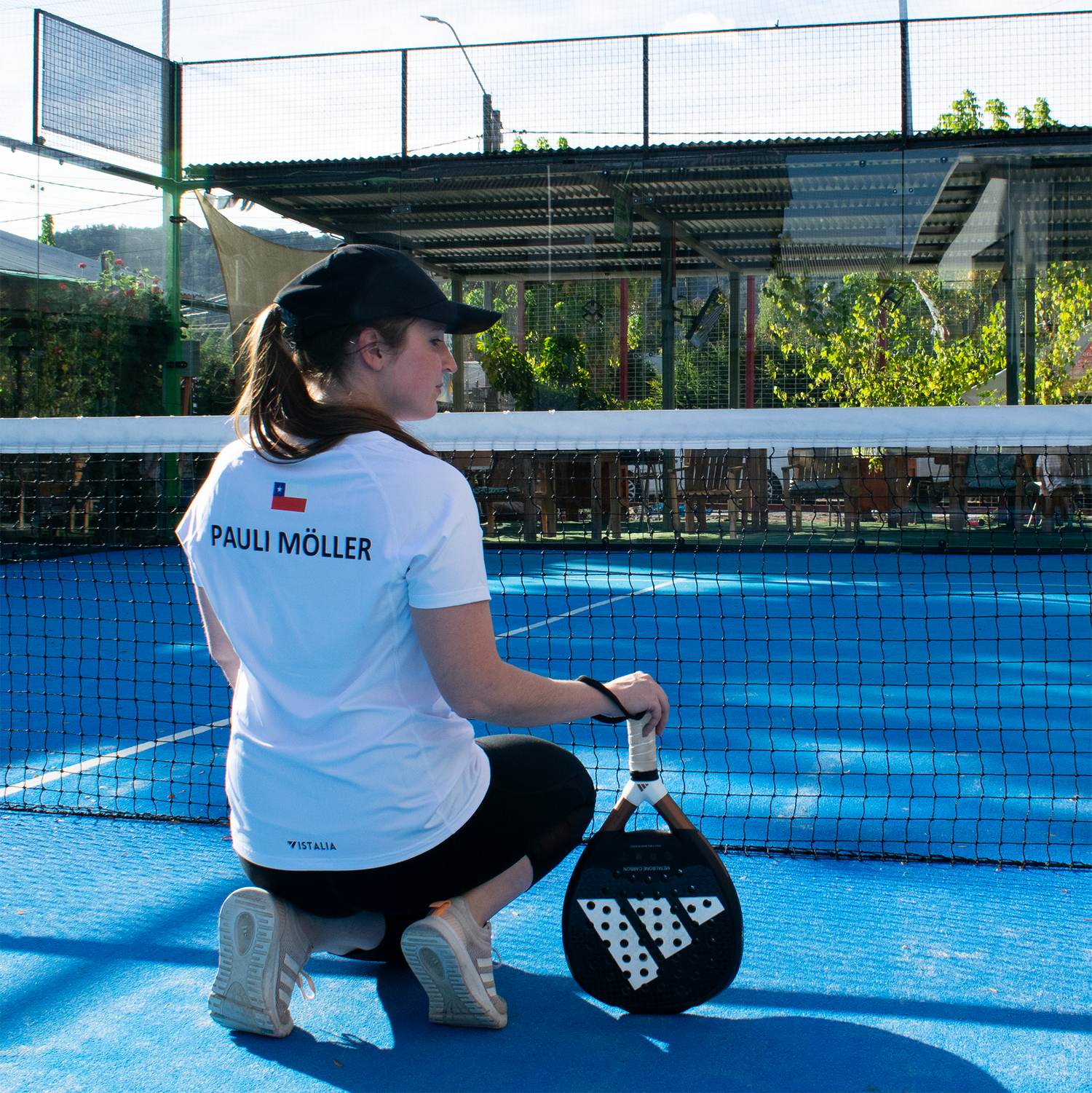 mujer en cancha de pádel de espaldas y cuclillas apoyando pala en el suelo