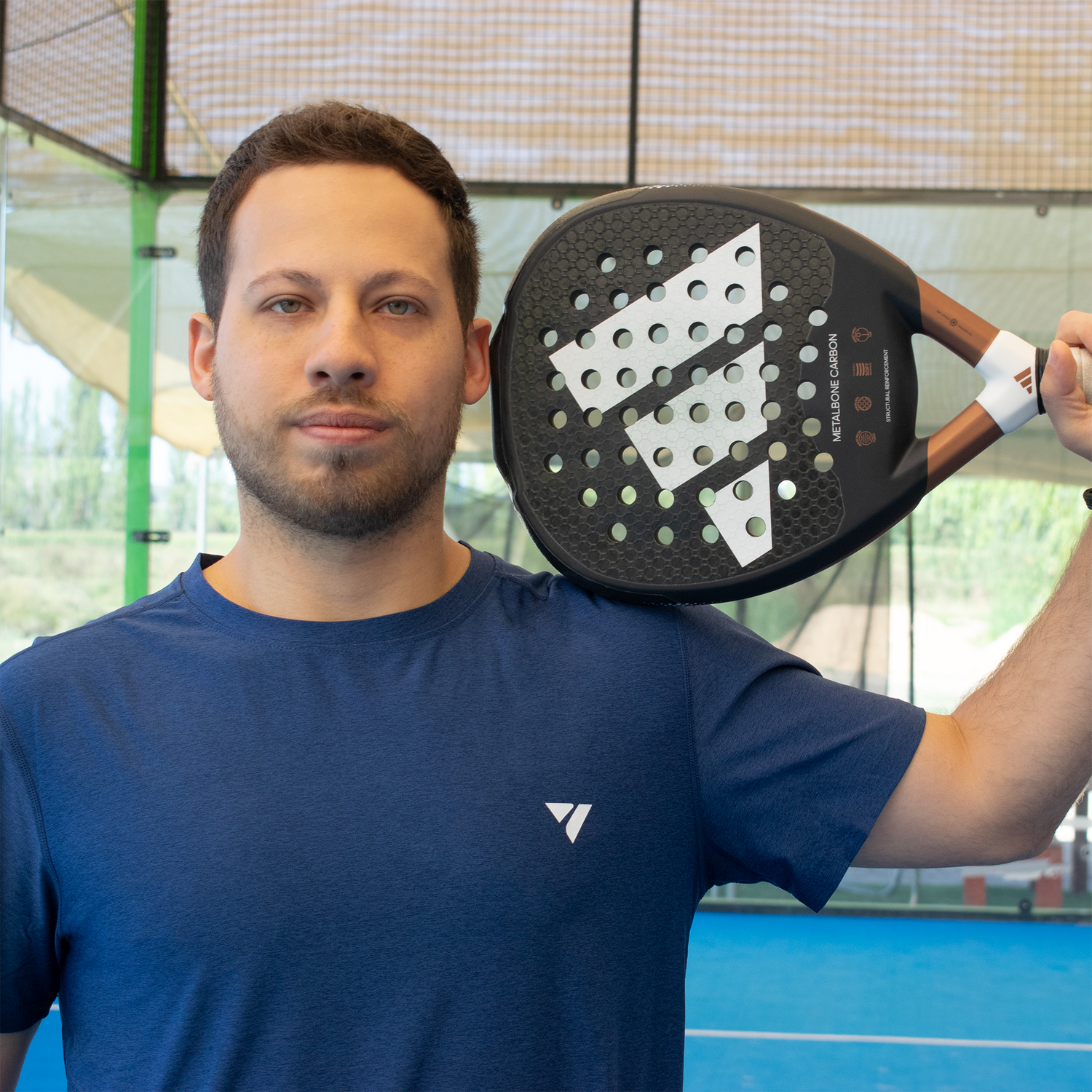 modelo usando polera azul con logo blanco en el pecho en cancha de pádel con pala en la cabeza