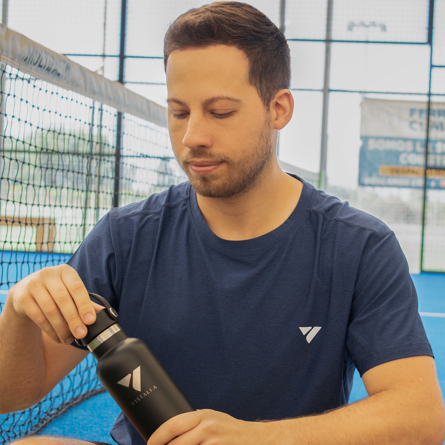 jugador de padel abriendo botella térmica Vistalia sentado sobre cancha de pádel