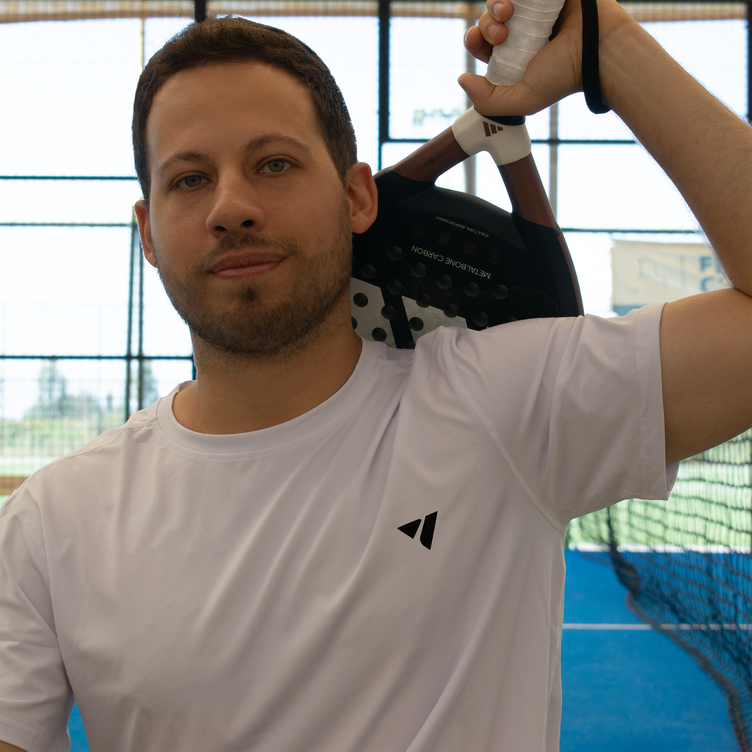 modelo con polera blanca y pala de pádel en la espalda sentado delante de cancha